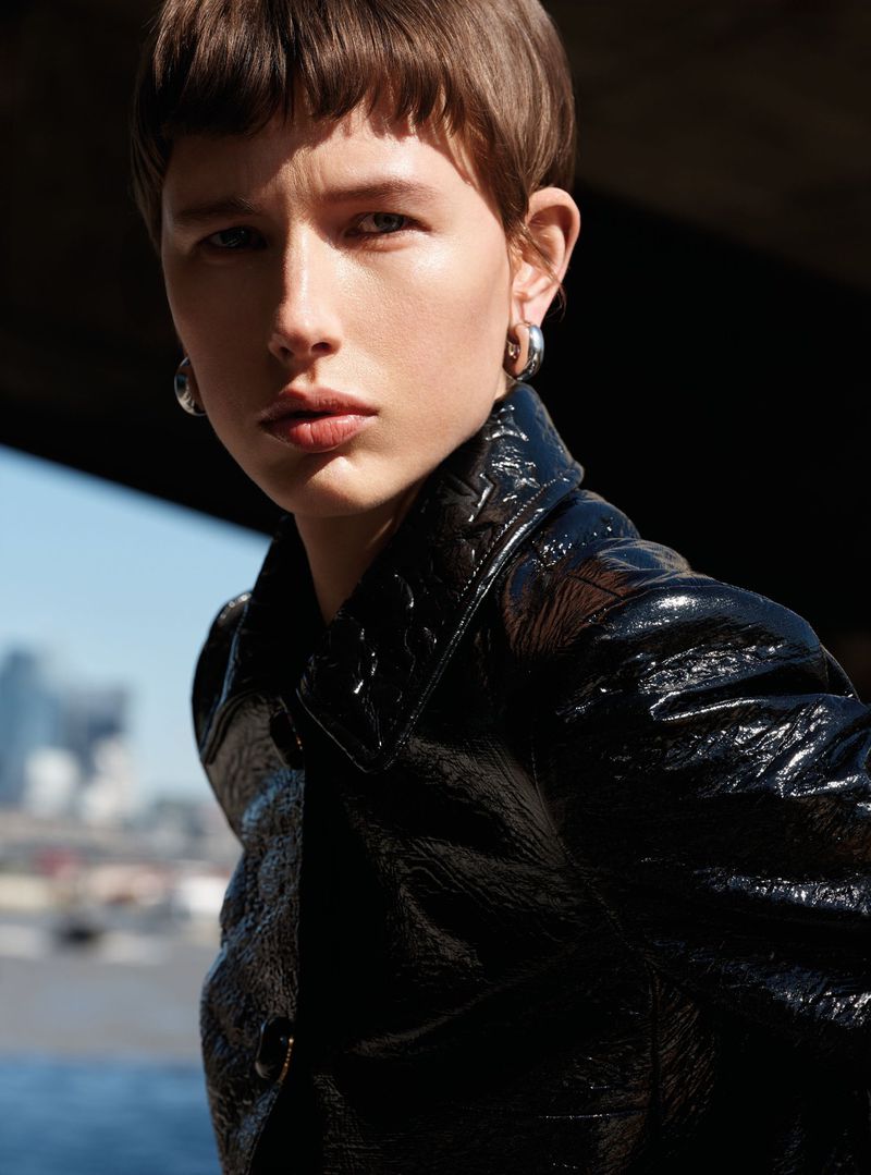 Model with short brown hair wearing a glossy black jacket and silver hoop earrings against an urban backdrop.