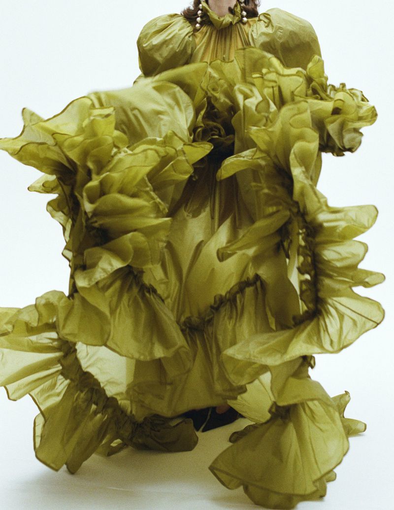 Model wearing an voluminous olive green ruffled gown against a plain white studio backdrop.