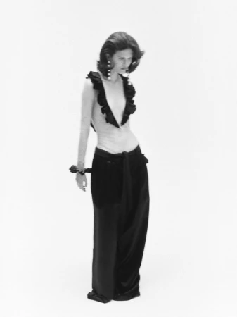 Monochrome fashion editorial image featuring a model wearing a ruffled top and dark trousers against a white background