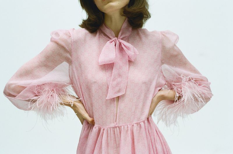 Model wearing a pink chiffon dress with a bow necktie and feather cuffs against a plain white background