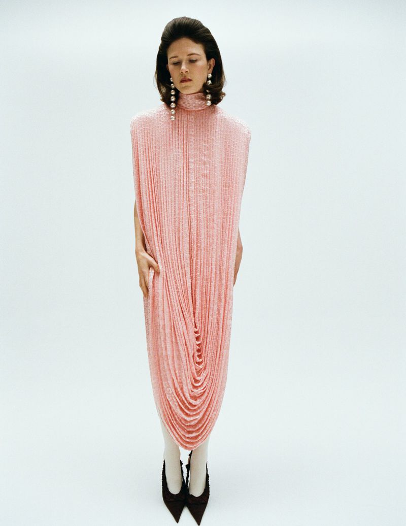 Model standing in a long pink textured high neck dress and black heels against a white background