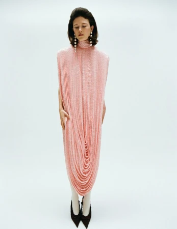 Model standing in a long pink textured high neck dress and black heels against a white background