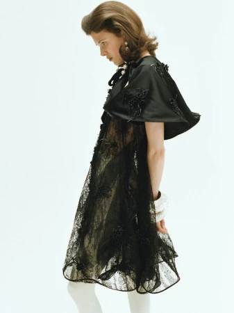 Model in black satin cape and lace dress standing against a plain white background