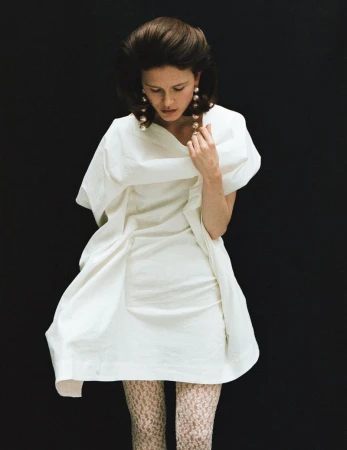 Model wearing a white dress and patterned tights with large pearl drop earrings against a solid black background.