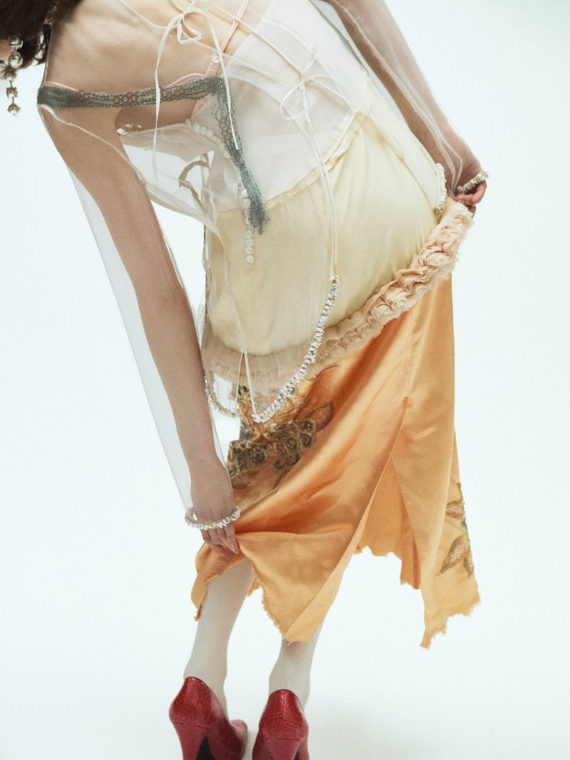 Model wearing layered sheer garments, an orange skirt, and red heels against a white studio background