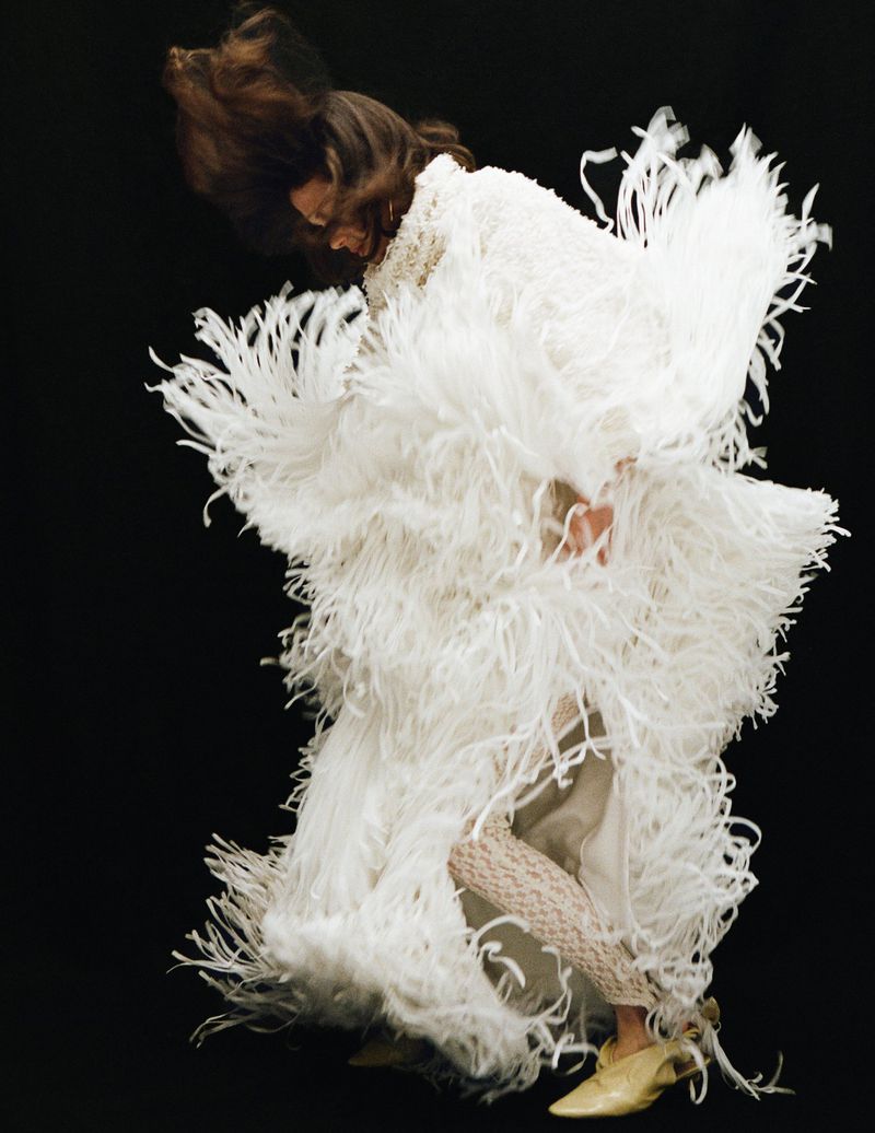 Model in motion wearing a heavy white fringed garment against a solid dark background