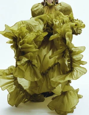 Model wearing an voluminous olive green ruffled gown against a plain white studio backdrop.