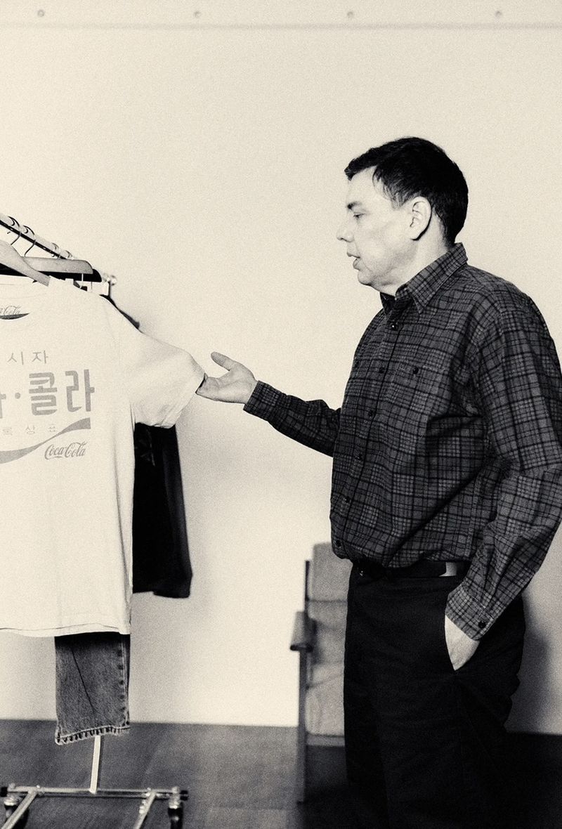 Monochrome portrait of an adult male standing next to a garment rack with a Coca-Cola t-shirt on display in an indoor setting.