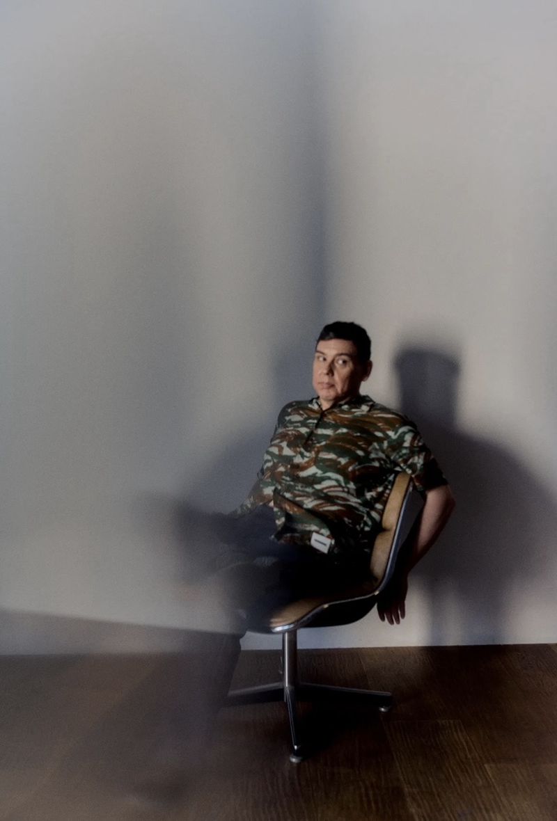 Portrait of an adult male sitting in a chair in front of a neutral wall. The man is wearing a patterned shirt.