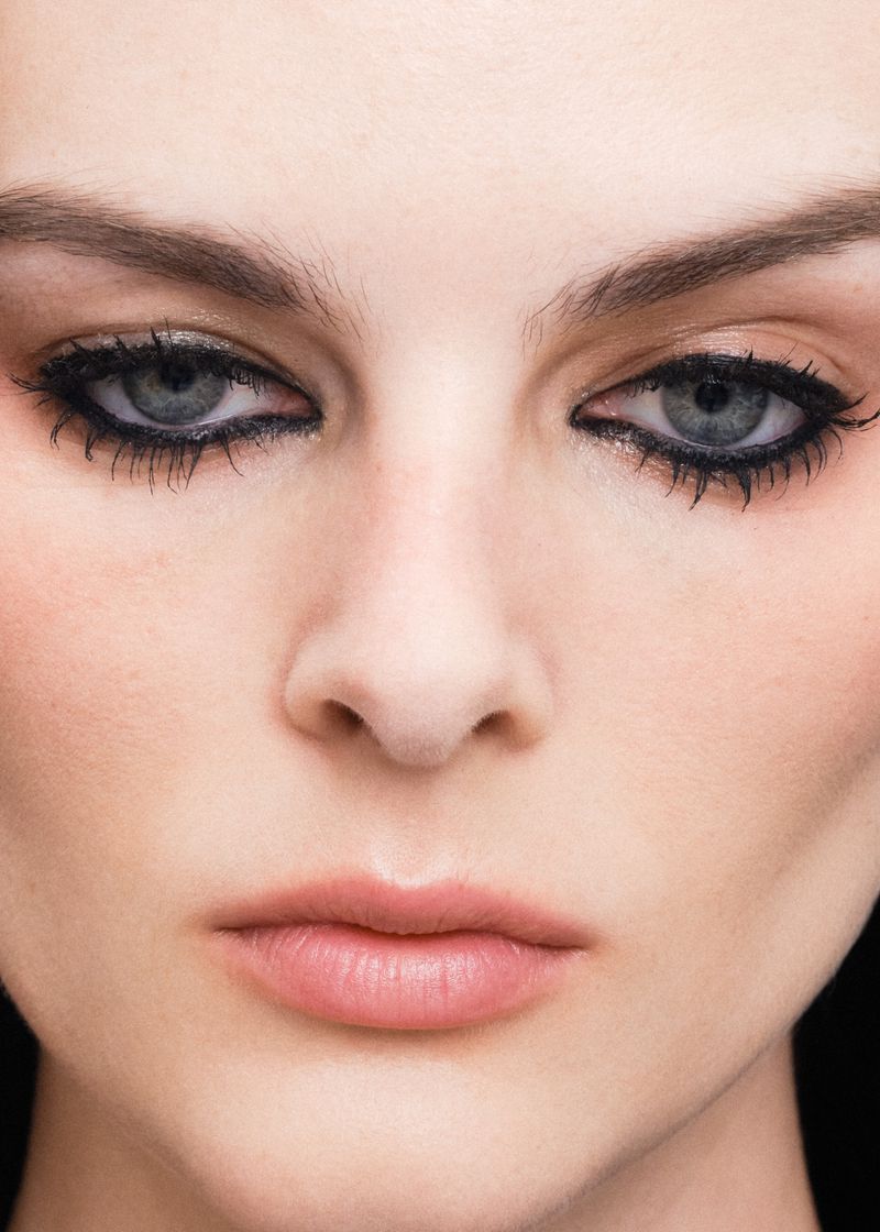 Close-up portrait of a woman with dark eyeliner highlighting her eyes. Features include fair skin, light pink lips, and neutral expression.