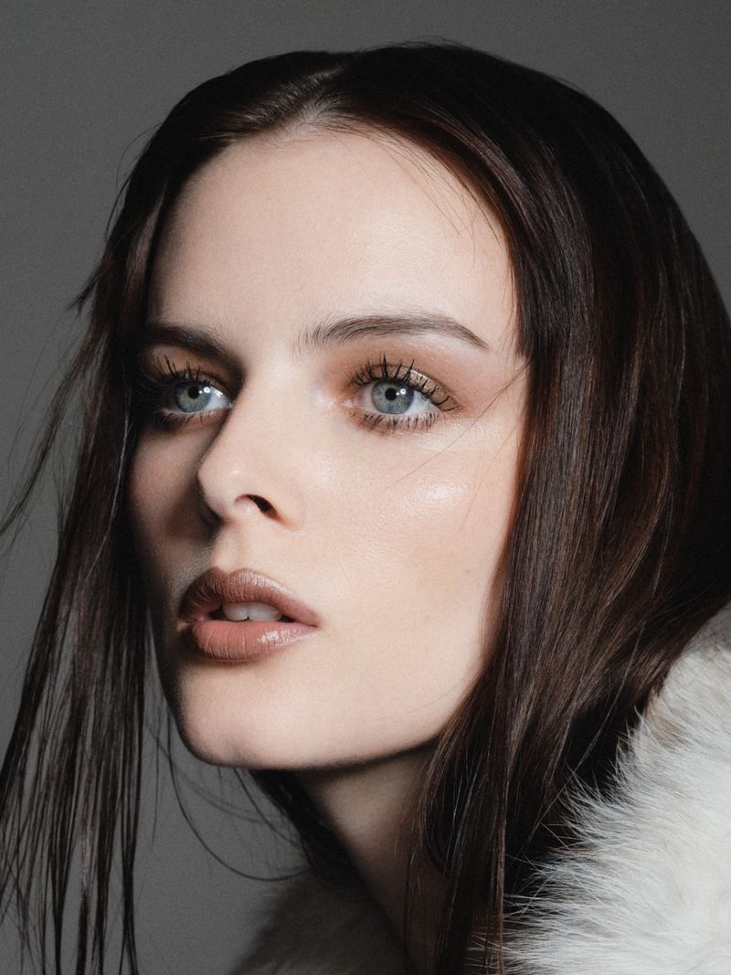 Portrait of a woman with fair skin, dark hair, and light blue eyes, posing indoors with a soft, diffused lighting and a grey background.