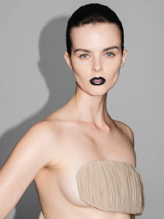 A nude portrait of an adult female model with wet hair, dark lipstick and a gathered light-brown fabric draped across her chest in a studio.