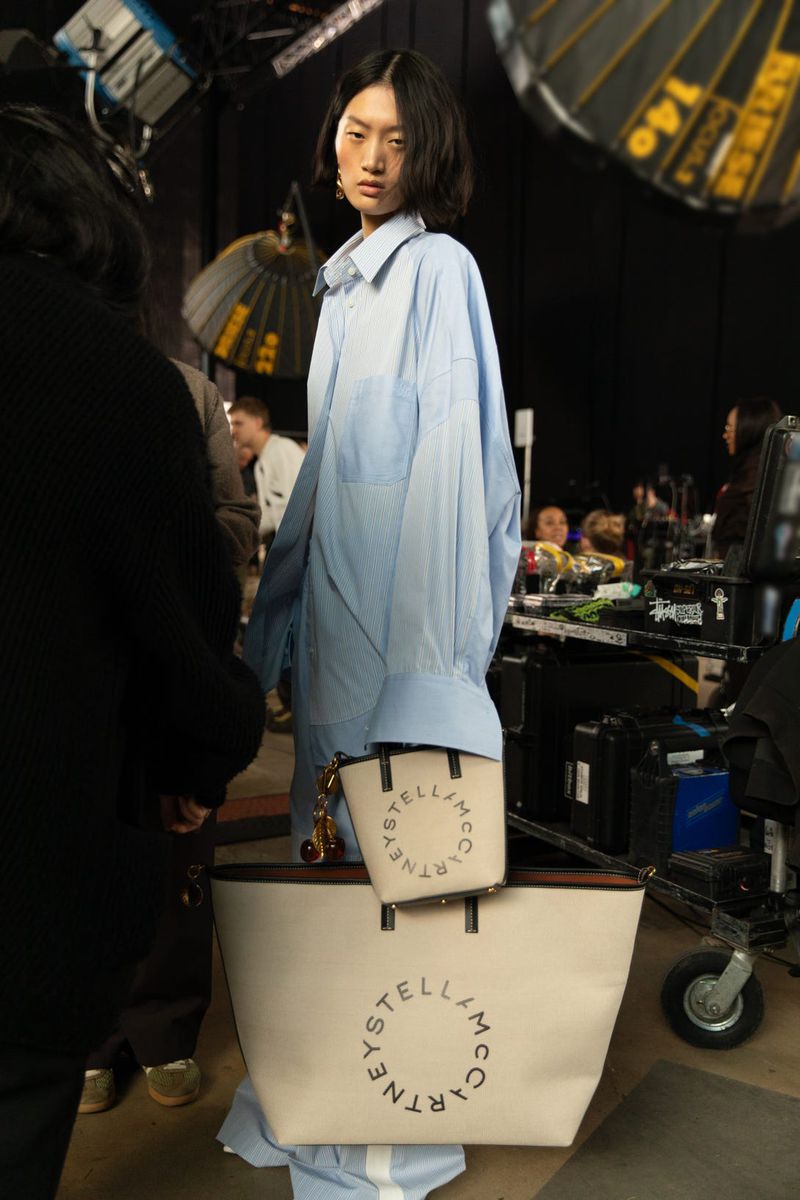 Model Yilan Hua wearing an oversized light blue shirt holding branded tote bags backstage at H&M x Stella McCartney 2026 lookbook shoot