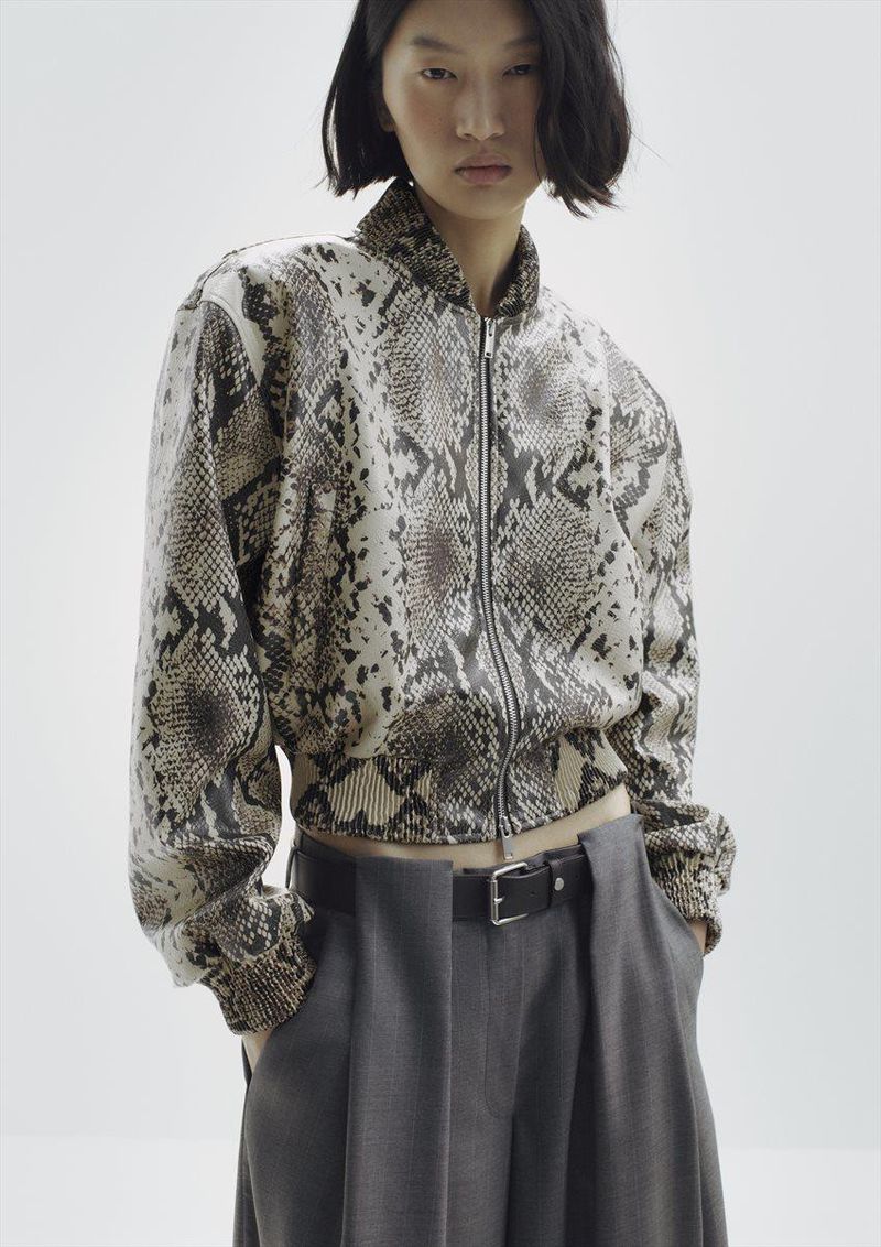 Model wearing a snakeskin print zip-up bomber jacket and grey trousers against a white background
