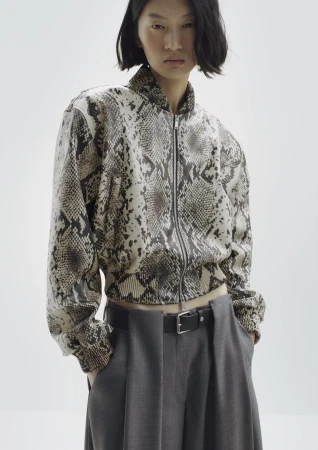 Model wearing a snakeskin print zip-up bomber jacket and grey trousers against a white background