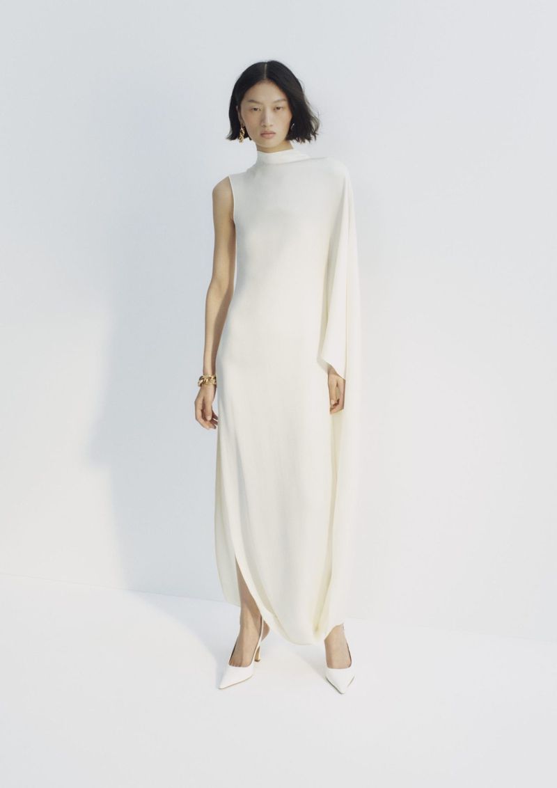 Model wearing a long white sleeveless dress and white heels against a seamless white studio background.