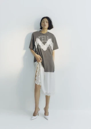 Model wearing a grey t-shirt and white lace skirt standing against a white studio wall