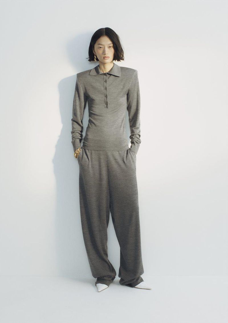 An Asian woman poses in a gray collared top and baggy pants against a white backdrop, showcasing a minimalist and fashionable look.