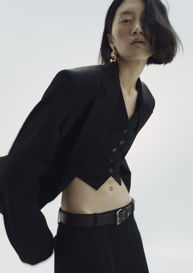 Model wearing a structured black blazer, vest, and trousers against a pale grey seamless background