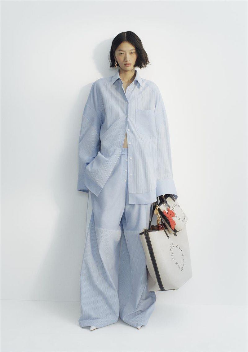 Model in oversized light blue striped shirt and matching trousers holding white tote bags against white background