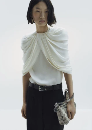 Model in white draped blouse and black trousers holding a snakeskin handbag against a plain background