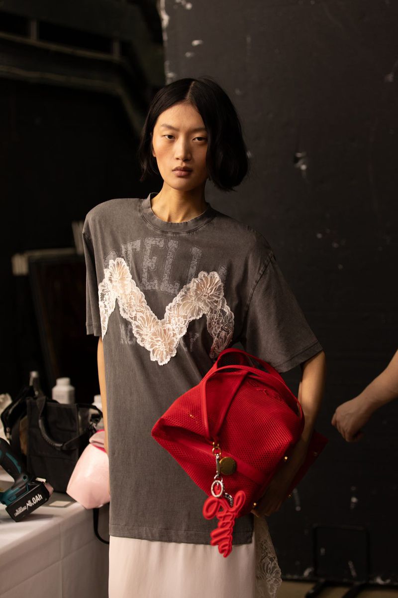 Model wearing dark grey graphic t-shirt and white skirt holding a bright red bag backstage for H&M