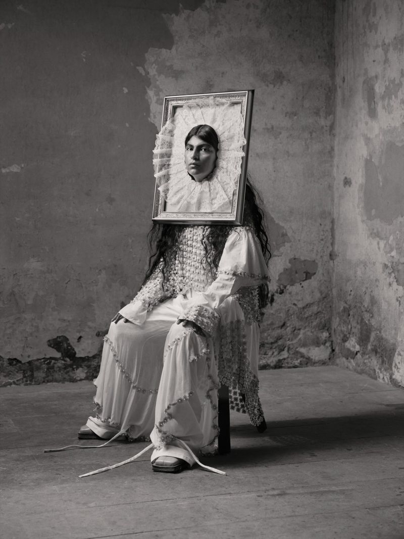 Monochrome portrait of a seated person holding an ornate framed picture framing their face with lace ruff against a rustic wall