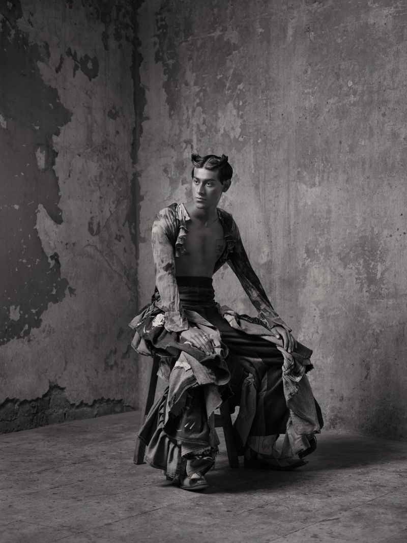 Portrait of a man seated on a stool wearing ruffled garments against a textured concrete wall in black and white