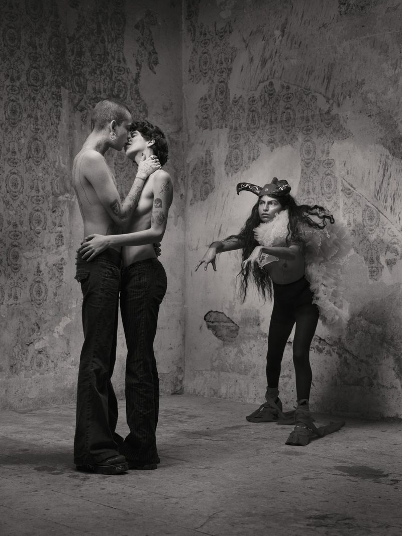 Two shirtless people embracing and kissing in a room with an ornamental wall; a third person in a feathered costume watches them. 
