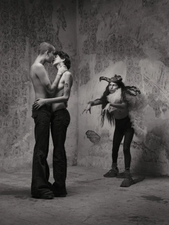Two shirtless people embracing and kissing in a room with an ornamental wall; a third person in a feathered costume watches them. 