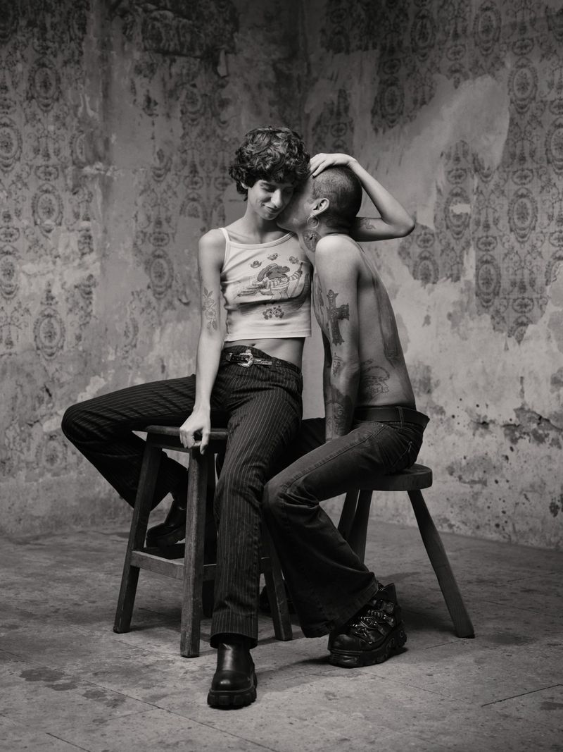 Two people seated on wooden stools against a weathered patterned wall in a black and white portrait photograph.