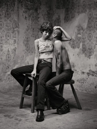 Two people seated on wooden stools against a weathered patterned wall in a black and white portrait photograph.
