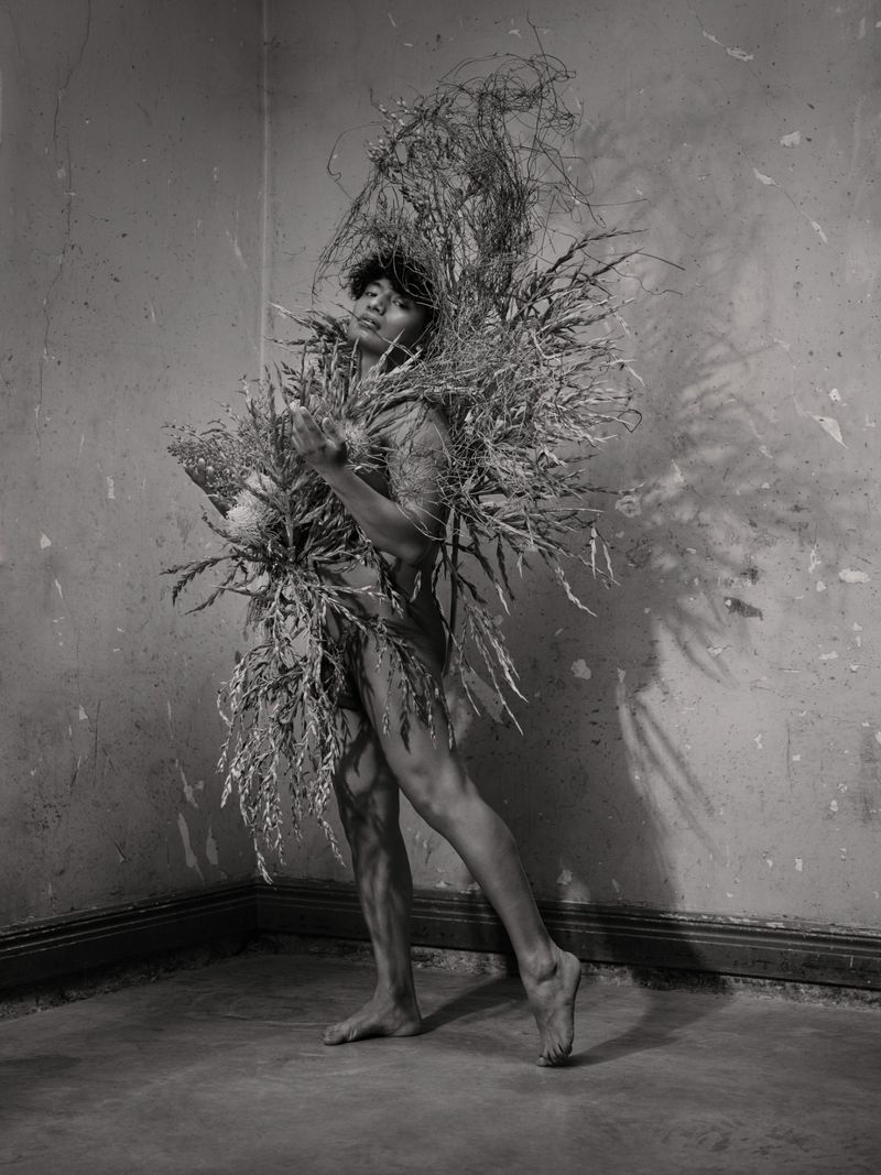 Monochrome portrait of a person adorned in elaborate dried botanical sculptures against a textured wall.