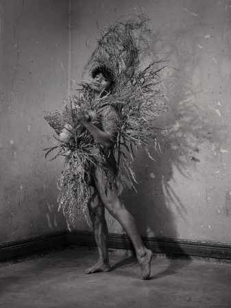 Monochrome portrait of a person adorned in elaborate dried botanical sculptures against a textured wall.
