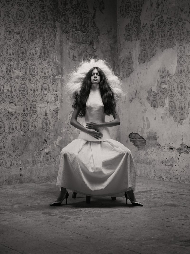 Monochrome portrait of a seated woman wearing a voluminous white dress and structured headpiece against a textured, patterned wall.