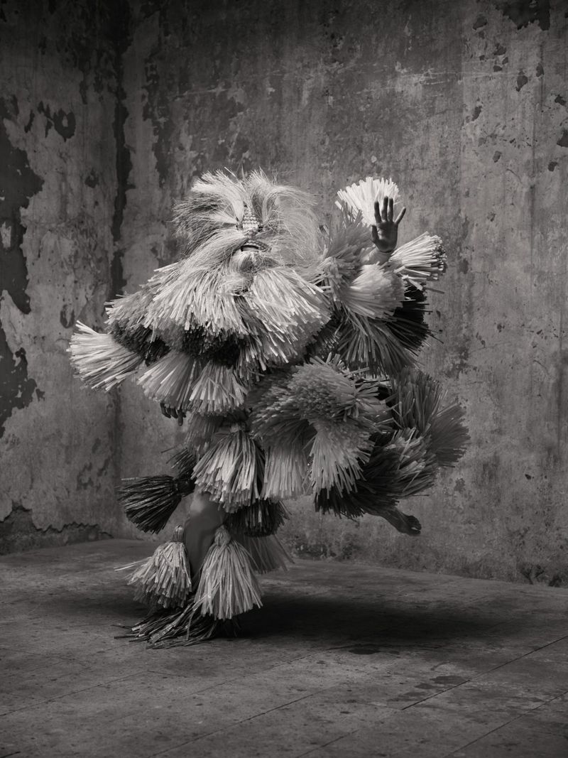 Person in fringed costume striking pose in front of weathered wall, monochrome image. Art, fashion, and performance.