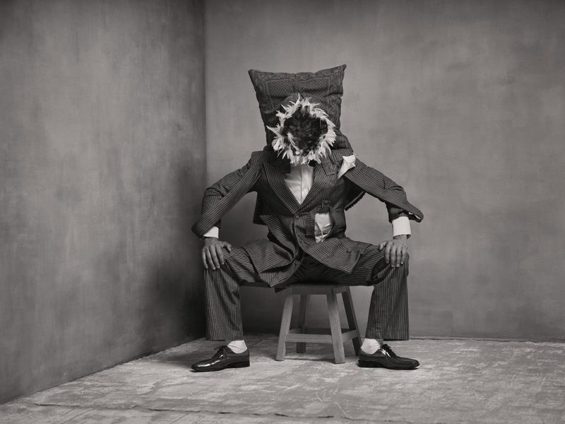Surreal portrait of a seated person in a structured suit with a feathered face and pillow head