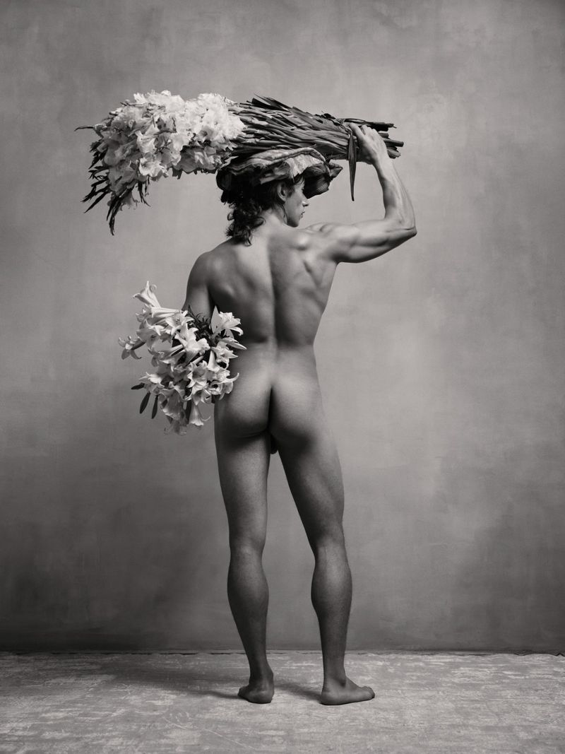 Monochrome portrait of a nude male figure carrying large bundles of white flowers on his head and arm.