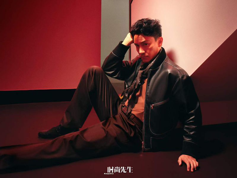 Asian man in a black jacket poses seated on the floor with a red background, looking sideways at the camera in a studio portrait.