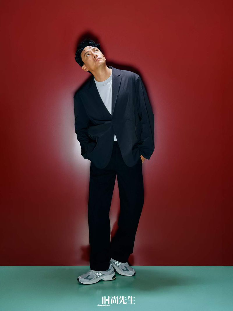 Man wearing a dark suit jacket, white t-shirt, and sneakers against a deep red studio background with teal floor.