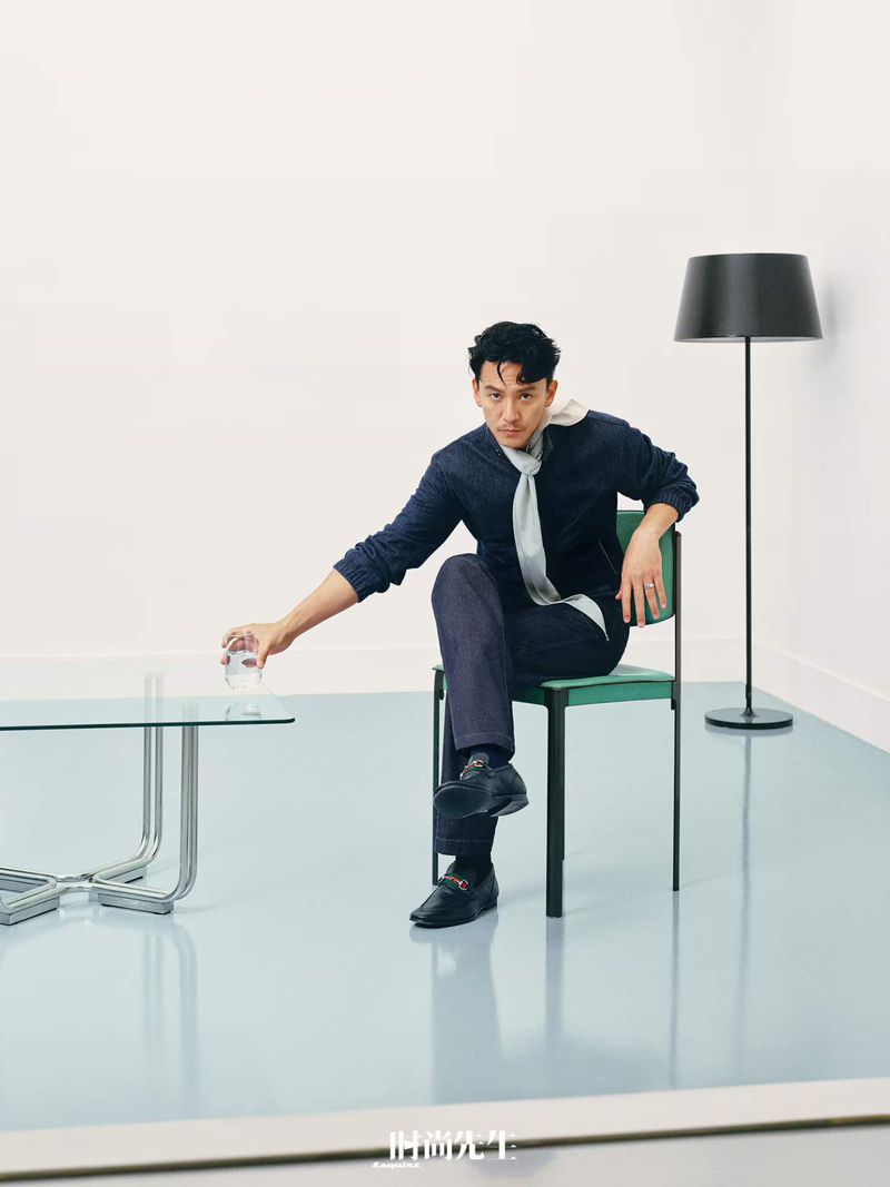 Man seated on a green chair holding a glass of water next to a glass table with a black floor lamp in the background.