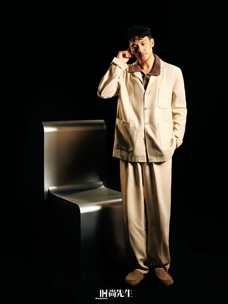Man wearing a cream jacket and trousers standing next to a metallic chair against a solid black background.