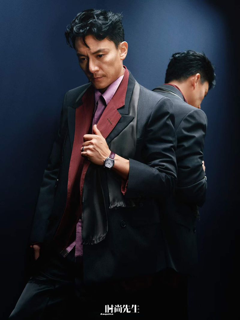 Man wearing a tailored dark suit jacket, maroon layer, and purple shirt with a wristwatch against a dark background