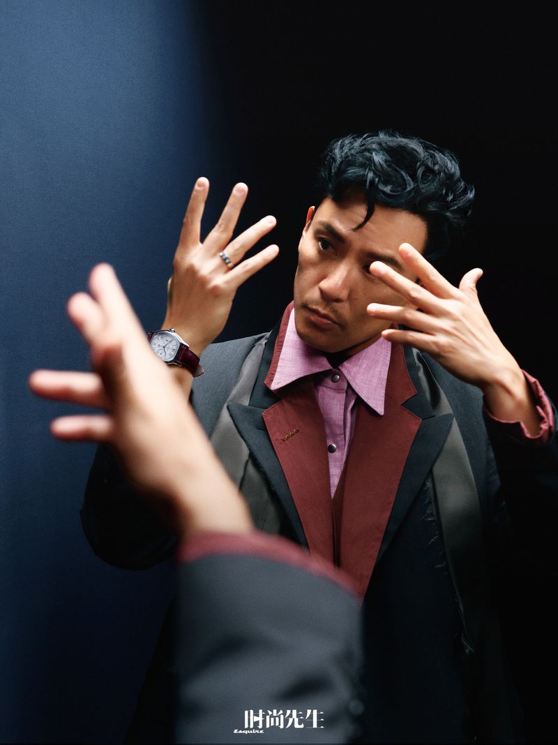 Man wearing dark suit with burgundy lapels and pink shirt posing with hands raised in front of dark background for Esquire China