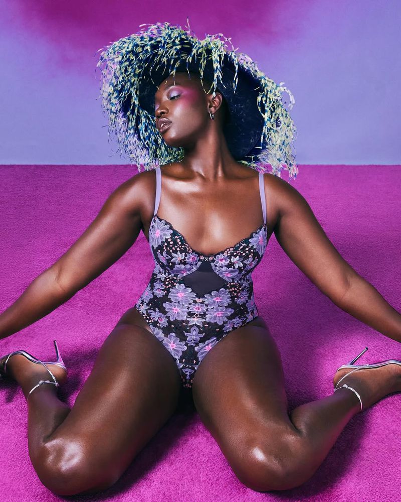 Model wearing purple floral lingerie and a large feathered hat kneeling on a pink carpet.