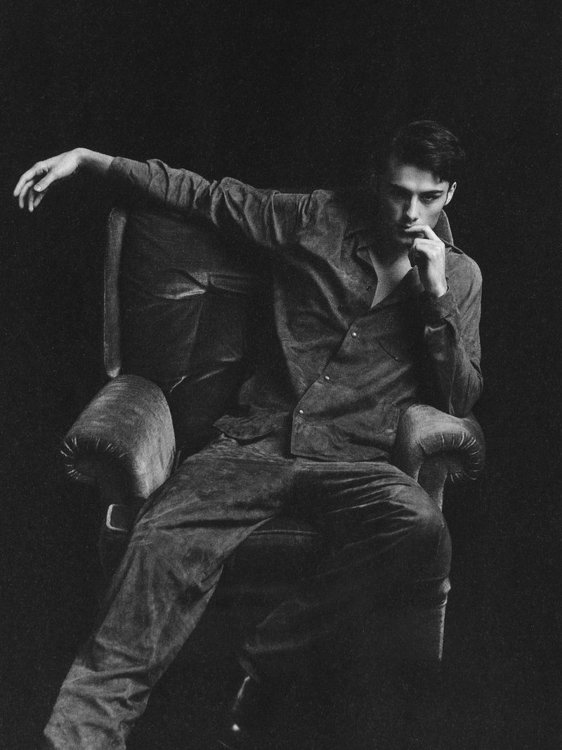 Monochrome portrait of an adult male in an open-collared shirt and trousers, seated in a wingback chair against a dark background.