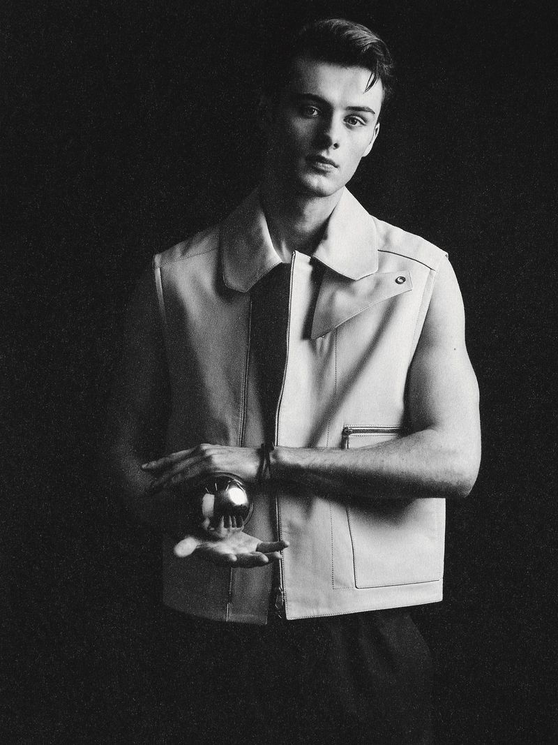 Monochrome portrait of a young adult man wearing a vest and holding a silver ball, set against a dark background. Fashion concept.