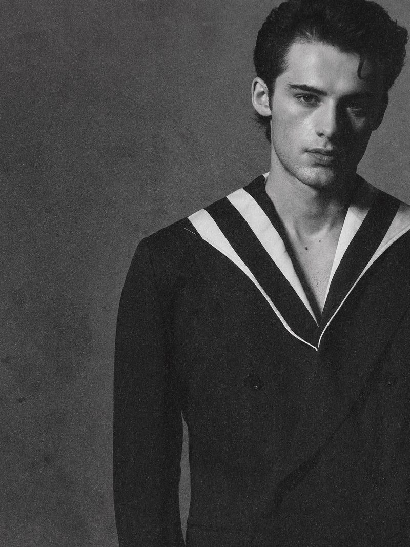 Black and white portrait of a young adult male wearing a dark double-breasted blazer with white striped lapels, looking at the camera.