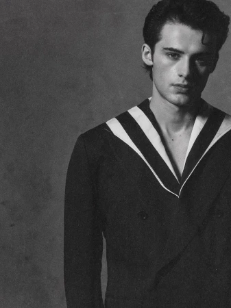 Black and white portrait of a young adult male wearing a dark double-breasted blazer with white striped lapels, looking at the camera.