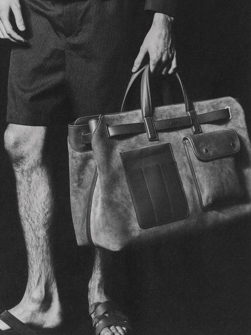 Monochrome studio shot of a man wearing shorts and sandals, holding a tan suede leather duffel bag against a dark background.